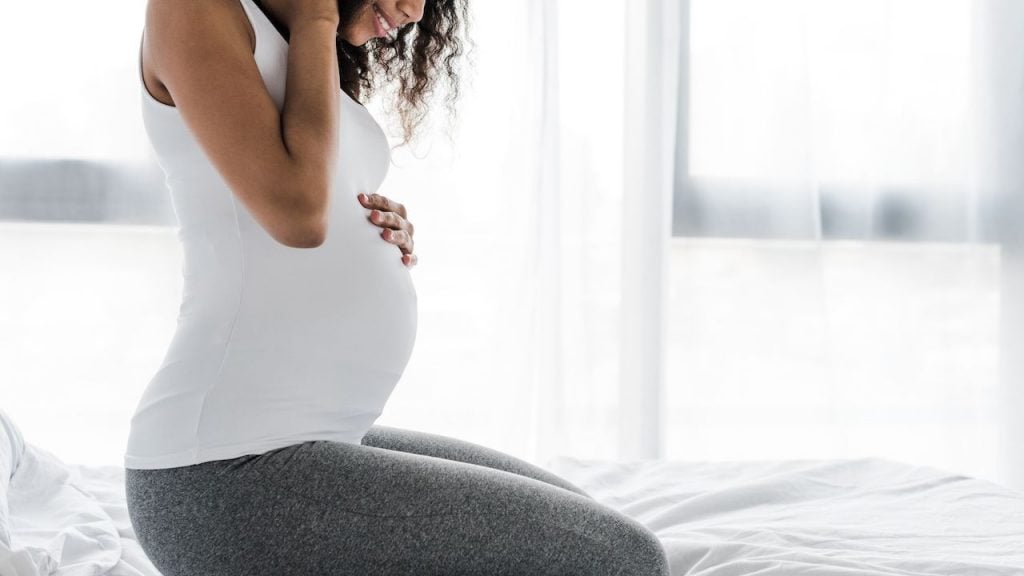 Cropped view of happy curly pregnant african american woman touching belly while sitting on bed — Photo